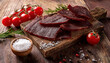 © Arda ALTAY - Sliced beef jerky is arranged on a rustic wooden board alongside fresh cherry tomatoes, rosemary, and a bowl of sea salt.