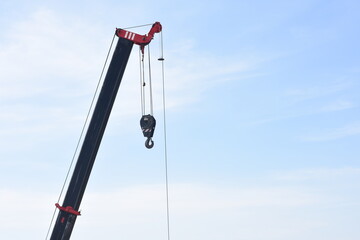 the upper part of a crane against a clear blue sky. The crane's arm is extended diagonally, and a hook is hanging from the end of the crane's cable.