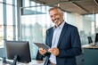 © NISHAN'S STUDIO - Happy middle aged business man ceo wearing suit standing in office using digital tablet. Smiling mature businessman professional executive manager looking away thinking working on tech device