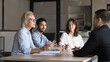 © fizkes - Group of four professionals sit around conference table engaged in discussion meeting event for decision-making, strategy planning, corporate goals sharing, led by mature businesswoman boss or coach