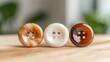 © familymedia - A close-up photo of three assorted buttons in different colors and designs, well-arranged on a wooden surface, showcasing detailed textures and craftsmanship.