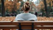 © familymedia - In a park during autumn, a young woman with a bun hairstyle sits on a bench among fallen leaves, highlighting a serene moment of solitude and nature's beauty.