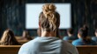 © familymedia - A man with a man bun hairstyle is seated in a classroom during a lecture. The room is dimly lit, with other students sitting in wooden desks around him.