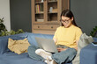 © Clique Images - Woman in eyeglasses and casualwear pressing keys of laptop keypad while sitting on sofa and crossing her legs