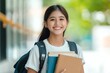 © Tondone - A cheerful student with long hair and a backpack smiles while holding books outdoors