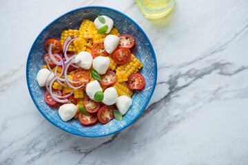 Wall Mural - Blue bowl with cherry tomato, mini mozzarella and corn salad, top view on a white stone background, horizontal shot with space