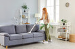 © Studio Romantic - Positive woman or maid, performing household chores, vacuuming a sofa during cleaning at home living room. Housecleaning and home service, emphasizing role in maintaining a clean household.