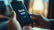 © PRDOM - A person pointing at a phone displaying a 'Book Now' button, suggesting an online booking action.