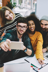 Fototapete - Multicultural group of successful students in stylish casual apparel making selfie photos on modern smartphone after good teamwork on common design project sitting at netbook in university