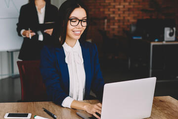 Fototapete - Positive smiling female finance director in stylish eyeglasses searching information via laptop computer while employees communicated on blurred background, concept of business and partnership