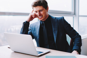 Fototapete - Cheerful male businessman in formal wear sitting at desktop and watching funny comedy video via multimedia application on modern laptop device, smiling male entrepreneur reading positive news