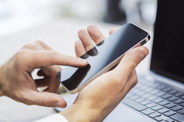 Poster - Man engaged with his modern smartphone, laptop slightly out of focus nearby against the blurred office hustle and bustle
