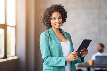 Leinwandbilder - A professional woman stands confidently in an office, holding a tablet. She smiles while engaging with her colleagues, highlighting teamwork and diversity in a corporate environment.