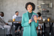 © Prostock-studio - A woman in a green suit stands confidently in a modern office, holding a tablet. Behind her, a diverse group engages in teamwork and discussion around a table, focused on their tasks.