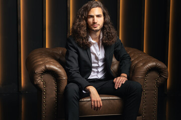Aufkleber - A confident European man with long hair is seated in a stylish armchair, dressed in formal business attire. He radiates professionalism and leadership in a modern office setting.