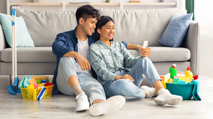 Wall Mural - Cute asian couple with cellphone having break after cleaning new apartment, looking for house-keeping service on Internet, young man and woman sitting on floor among cleaning tools, home interior