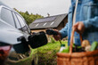 © Halfpoint - Close up of man holding basket with groceries, plugging charger into an eletric car. House with solar panel system on roof behind.