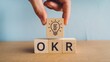 © Naruedetch - Close up of a hand holding a light bulb icon over wooden blocks with the text OKR signifying objectives key results and goals  The image represents the concept of innovation creativity