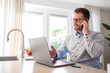© erikrous - Working at home in the kitchen. Caucasian male sitting at the kitchen counter behind his laptop working and communicating. Wearing a light blue formal shirt and glasses