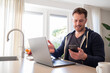 © erikrous - working at home in the kitchen. caucasian male sitting at the kitchen counter behind his laptop working and communicating. Dressed in a hoodie, holding phone in the hand on a video call.