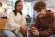 © Studio Marmellata - A woman in a beige sweater sitting next to a man wearing a brown hoodie indoors, pointing at his smartphone and discussing something in a casual, friendly setting