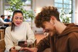 © Studio Marmellata - A woman in a beige sweater holding a smartphone and a young man in a brown hoodie laughing and talking together in a comfortable indoor cafe with a lively, well-lit environment