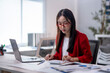 © Apichat - A woman in a red jacket is sitting at a desk with a laptop and a calculator