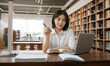 © RooftopStudioBangkok - focused Asian university student studies in library, surrounded by books. She holds pen and looks at camera, conveying determination and engagement in her studies