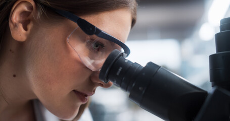 Fototapete - Close Up Portrait of a Female Microbiologist Wearing Safety Glasses and Looking Through the Microscope, Analyzing Sample Cells. Skillful Scientist Working in a Medical Science Laboratory