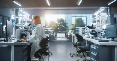 Leinwandbilder - Medical Research and Development Laboratory: Beautiful Caucasian Female Scientist Working on a Computer and Looking at a Petri Dish Sample Under a Microscope. Advanced Biotechnology Lab