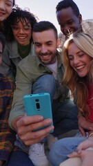 Wall Mural - Vertical HD video of happy group of young people having fun sharing video content on social media platform. Millennial multiracial friends using smartphone while sitting outdoors