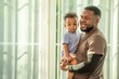 © BESTIMAGE - Happy african black parents dad father throw baby son on piggyback and neck riding teasing fun near window. Black baby son and daddy enjoy teasing kiss cheek and neck riding at window light