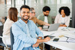© Vadim Pastuh - A young man in a blue shirt smiles confidently at the camera, with his colleagues engaged in a discussion in the background, showcasing a harmonious team in a productive setting