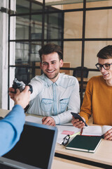 Poster - Successful male colleagues enjoying work time together sitting at desktop indoors while one of them messaging via smartphone, smiling hipster guy taking retro camera from friend on front with laptop