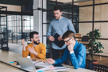 Fototapete - Group of happy male coworkers laughing and having fun during cooperation and research using netbook and wifi, positive designers creating sketches together on big wooden table in coworking space