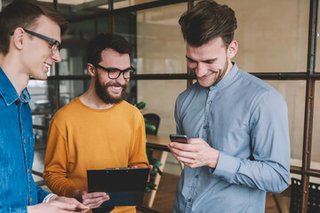 Poster - Successful male partners enjoying break of work and laughing from funny joke reading in social network via smartphone device, young positive men communicated and using technology in modern office