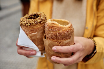 Close-up of woman holding two chimney cakes wrapped in paper with blurred city in background. Tourist enjoying freshly baked trdelnik at street in Prague, Czech Republic