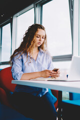 Fototapete - Concentrated millennial hipster girl chatting with colleague via telephone device connected to free wireless internet, serious female checking notification on cellular sitting with laptop indoors