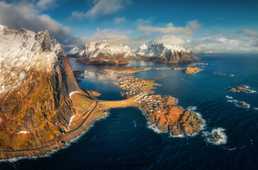 Wall Mural - Panoramic aerial view of Reine fishing village at sunrise. Winter in Lofoten islands, Norway. Colorful top drone view of snowy mountains, sea, road, reflection in water, blue sky with clouds at dawn