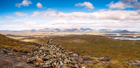 Wall Mural - Pile of rocks on a hill of a mountain, vast empty area and mountains in the background under blue cloudy sky. Connemara, county Galway, Ireland. Stunning Irish nature view on Twelve Bens mountains.