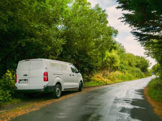 Wall Mural - A white medium size van is parked on a small county road in rural area with a red tail light. The road is empty and the sun is shining brightly. Product delivery to remote locations concept.