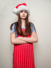 Wall Mural - Young teenager girl wearing blue shirt, Santa Claus hat and red and white apron on light color background. Cooking for special event concept. Model with long high volume hair and slim body type.