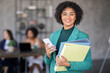 © Prostock-studio - A confident businesswoman stands in a modern office, holding folders and a smartphone. Behind her, colleagues collaborate on a project, emphasizing teamwork and diversity in the workplace.