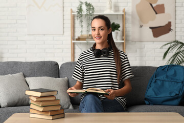 Wall Mural - Beautiful happy female student with stack of books, backpack and headphones sitting on sofa at home