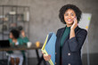 © Prostock-studio - A professional woman smiles while holding a folder and talking on her phone in a contemporary office. In the background, her colleagues are engaged in a meeting, showcasing teamwork.
