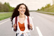© Prostock-studio - Joyful young lady in casual summer clothes walking along asphalt road in countryside, hitchhiking for ride outdoors. Lovely millennial woman with backpack traveling alone by autostop