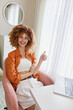 © SHOTPRIME STUDIO - Cheerful young woman in an orange shirt giving a thumbs up at her home office, representing positivity and productivity in a modern workspace