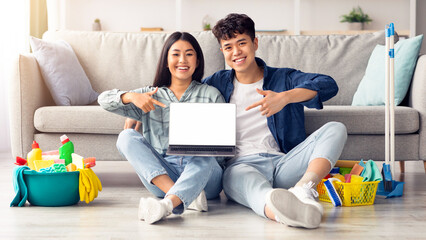 Wall Mural - Cheerful asian couple showing laptop with empty screen, smiling korean man and woman sitting on floor in living room, having break after cleaning home, recommending online house-keeping service