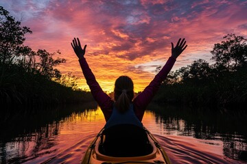 Fototapete - A person in a kayak celebrates a vibrant sunset over calm waters surrounded by greenery.