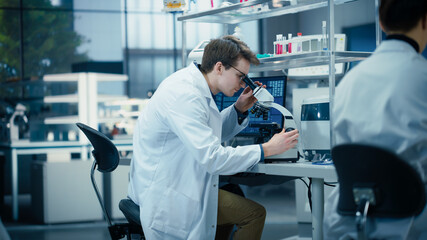 Leinwandbilder - Molecular Biology Research Scientist Looking at a DNA Sample Under a Microscope in an Applied Science Laboratory. Portrait of a Young Lab Engineer in White Coat Working on Vaccine Products